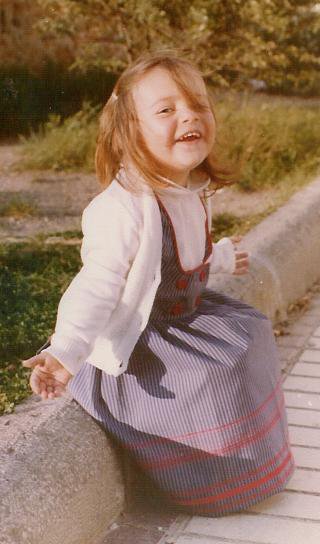 Niña pequeña, de 4 años, sentada en un bordillo ríe y abre sus brazos feliz.