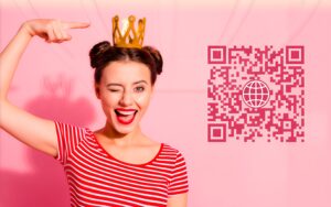 Chica sonriente, vestida con una camiseta de rayas fucsia y rosa, con el pelo recogido en dos moños laterales, ríe con la boca abierta, guiña su ojo derecho y señala con su dedo la corona dorada y pequeña que tiene en su cabeza. En el lado derecho de la chica hay un código QR fucsia, para escanear e ir a las redes sociales.